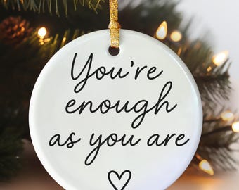 May include: A white ceramic Christmas bauble with the message "You're enough as you are" in black script, and a heart symbol. It hangs from a gold ribbon, with a blurred background of a Christmas tree and fairy lights.