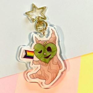 May include: A brown and pink cartoon character with a green heart-shaped face holding a rainbow flag. The character is attached to a gold star-shaped keychain.