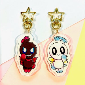 May include: Two acrylic keychains with gold star-shaped clasps. The keychain on the left features a brown and red cartoon character with a devilish expression. The keychain on the right features a white cartoon character with a halo and wings.