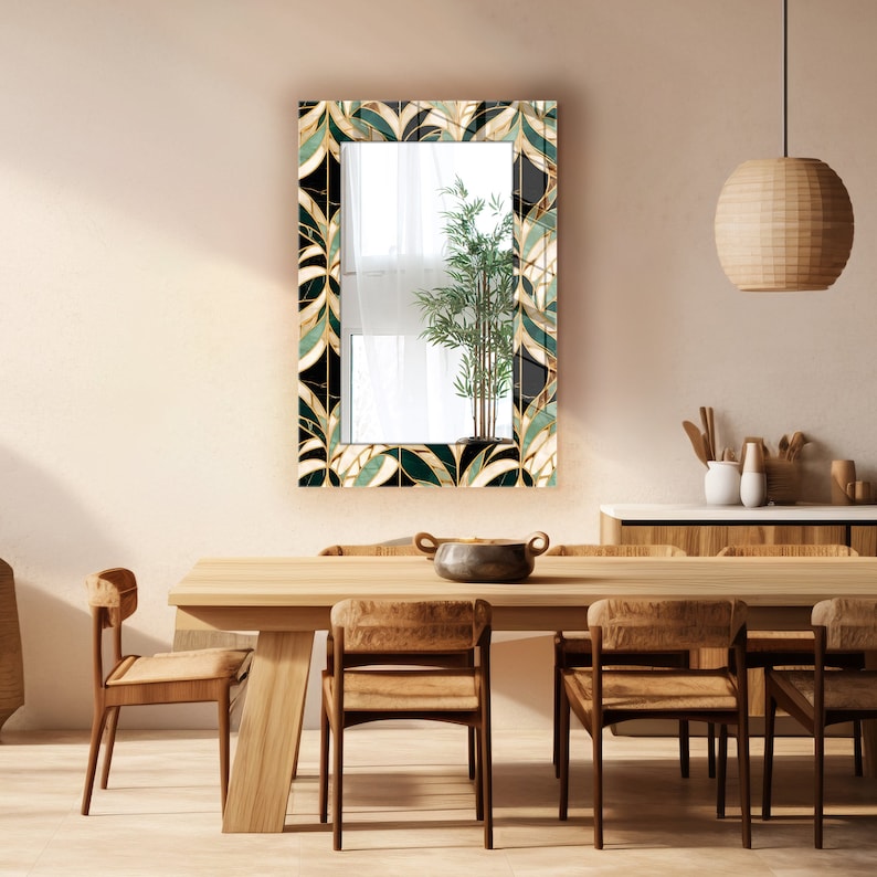May include: Rectangular wall mirror with a decorative border featuring a leaf design in shades of green, black, and gold. The mirror is mounted on a light-coloured wall above a wooden dining table with chairs. A bamboo plant is reflected in the mirror.