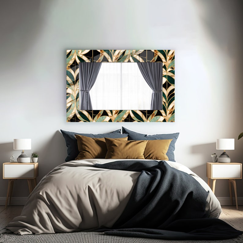 May include: A rectangular mirror with a decorative frame featuring a leaf pattern in gold, black, and green hangs above a bed. The bed has a beige duvet, blue pillows, and a dark blue throw. Two white bedside tables with lamps flank the bed.