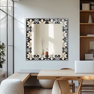 May include: A black framed mirror with a white floral design. The mirror is hanging on a white wall above a wooden table.