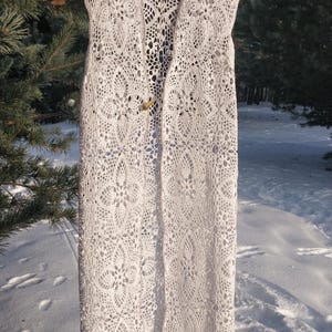 May include: A long, cream-colored, open-front vest with a delicate, openwork crochet design. The vest is sleeveless and hangs on a wooden hanger. A wooden button secures the front. The background shows snow and evergreen trees.