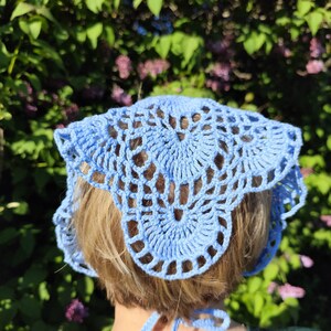 May include: A light blue, crocheted head covering with a scalloped edge. The head covering is made of a delicate, open-weave pattern. The item is tied at the back with matching blue ties. The background is a blurred view of green foliage and purple flowers.