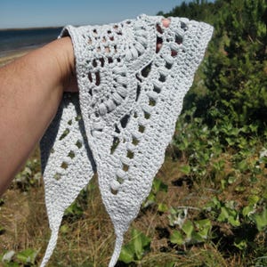 May include: A light gray crochet bandana featuring an intricate openwork pattern with textured puff stitches and a triangular shape. The accessory has long, thin crocheted ties at the corners and a scalloped edge design throughout the fabric.
