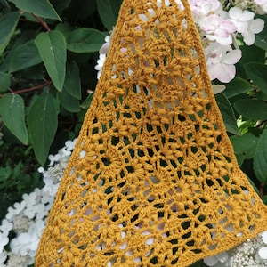 May include: Mustard yellow crochet bag with a floral pattern. The bag is triangular in shape and has a strap at the top. The bag is hanging in front of a background of white flowers and green leaves.