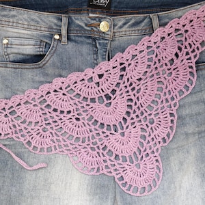 May include: A light purple triangular crochet scarf featuring an intricate openwork fan pattern with thin tie strings at the corners, layered over a pair of blue denim jeans with a metal button and silver-toned rivets visible at the waist.