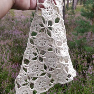 May include: Cream-colored, open-weave crochet piece with a floral pattern. The handmade item is held against a backdrop of green trees and purple flowers. The crochet work appears to be cotton or a similar natural fiber.