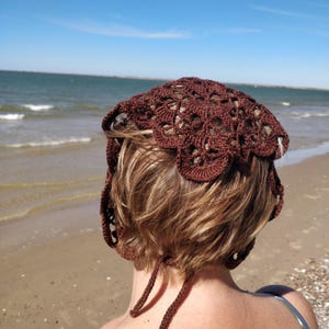 Puede incluir: Un sombrero marrón tejido a ganchillo con un borde festoneado y cuerdas para atar. El sombrero se lleva en la cabeza de una persona, con el océano y la playa al fondo. El diseño calado del sombrero permite vislumbrar el cabello que hay debajo.