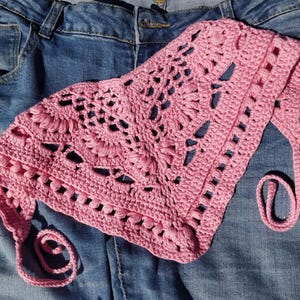 May include: A triangular crochet bandana in a solid light pink color. The accessory features an intricate openwork pattern with textured puff stitches and scalloped edges, finished with two long crocheted ties at the corners for securing.