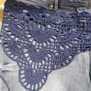 May include: A dark navy blue crochet shawl featuring an intricate fan-like pattern with openwork detailing. The garment is crafted with a series of interconnected circular motifs that create a scalloped edge, layered over a pair of blue denim jeans.