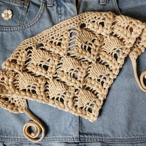 May include: A beige crocheted hat with a decorative floral pattern. The hat has a rounded shape and a curled, rope-like detail. The hat is made of a light-colored yarn and is set against a denim background.
