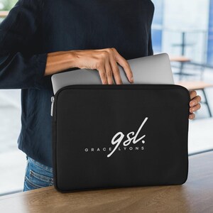 May include: Black laptop sleeve with the white text "gsl. GRACE LYONS". The sleeve is held by a person, with a silver laptop partially visible inside. The background is blurred, with a wooden table and a window.
