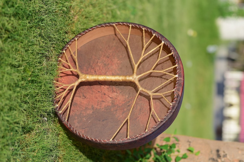 May include: A brown leather drum with a tree-like design made of woven rope. The drum is round and has a diameter of about 30 cm.
