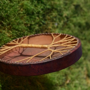 May include: A brown leather drum with a tree of life design made of woven rope. The drum is suspended in the air against a green mossy background.