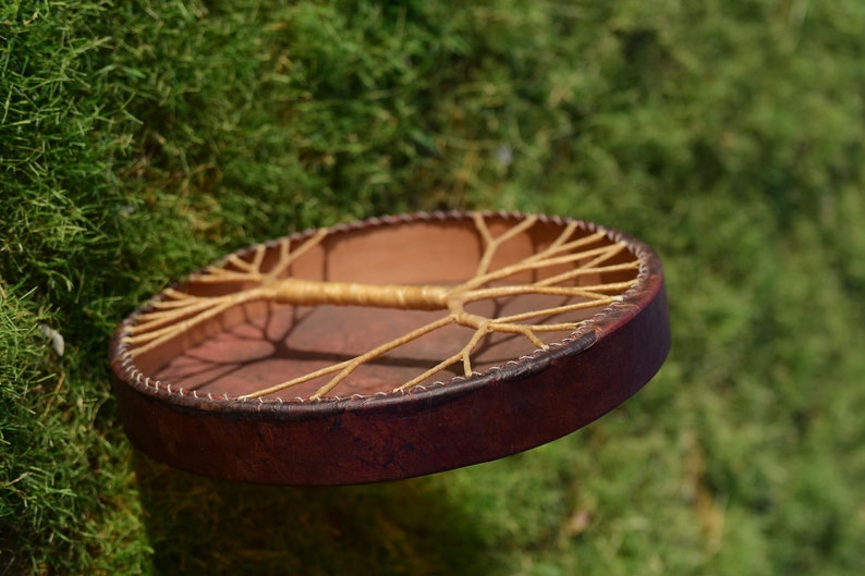 May include: A brown leather drum with a tree of life design made of woven rope. The drum is suspended in the air against a green mossy background.