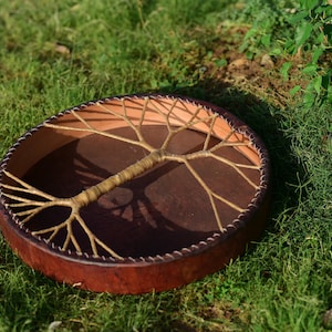 May include: A brown leather drum with a tree of life design made of woven rope. The drum is lying on a bed of green grass.