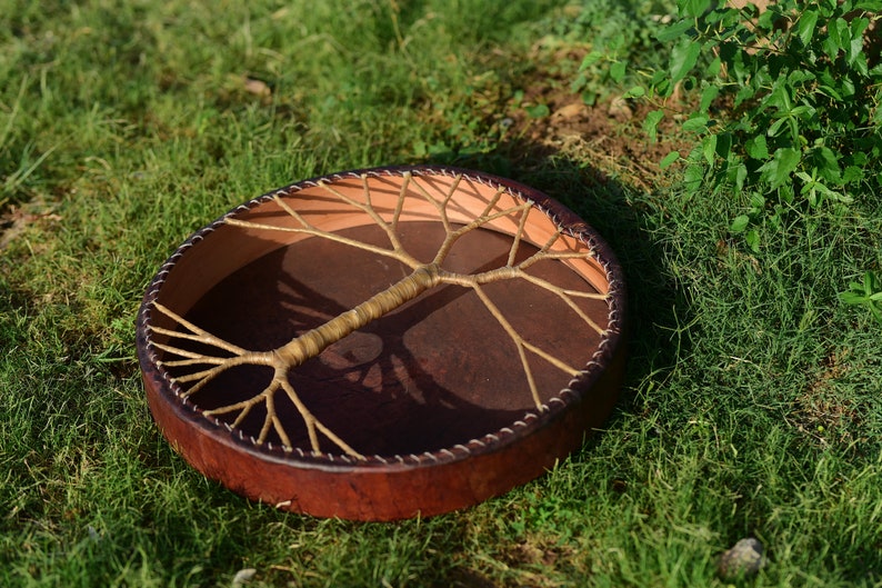 May include: A brown leather drum with a tree of life design made of woven rope. The drum is lying on a bed of green grass.
