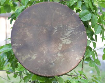 Pode incluir: Um tambor redondo, com aspeto antigo, com uma superfície castanha desgastada. O tambor está colocado contra um fundo de folhas e ramos verdes, sugerindo um ambiente ao ar livre. A superfície do tambor tem um aspeto texturizado.