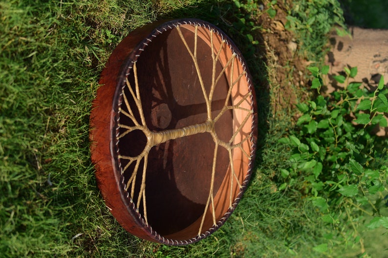 May include: A brown leather drum with a tree of life design made of woven rope. The drum is sitting on a bed of green grass.