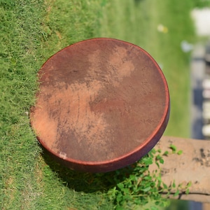 May include: A brown leather drum with a natural finish. The drum is sitting on a bed of green grass.