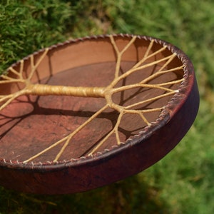 May include: A brown leather drum with a tree of life design made of woven rope. The drum is suspended in the air against a green grassy background.