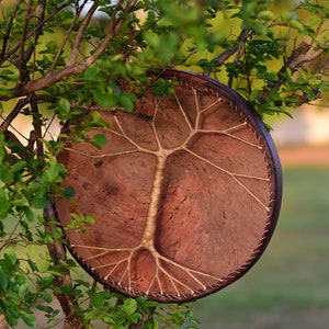Può includere: Un tamburo di pelle marrone con un disegno ad albero fatto di corda bianca. Il tamburo è appeso a un ramo d'albero.