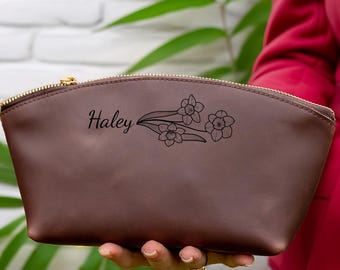 Könnte beinhalten: Eine dunkelbraune Kosmetiktasche aus Leder mit einem goldfarbenen Reißverschluss. Auf der Vorderseite ist der Name Haley in Schreibschrift eingraviert, ergänzt durch eine Strichzeichnung von drei Narzissenblüten.