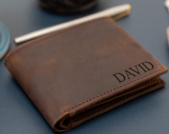 Könnte beinhalten: Eine braune Lederbrieftasche im Bifold-Design mit dem eingravierten Namen DAVID in einer dunklen Serifenschrift auf der Vorderseite. Die Brieftasche weist sichtbare Nähte an den Kanten und eine glatte, matte Textur auf, als einzelnes Accessoire.