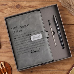 May include: A gray notebook with the text Difference Maker and Daniel printed on the cover, presented in a gift box with a black pen and a metal pen refill. The set includes a brown leather strap, a round brown leather holder with golf tees.