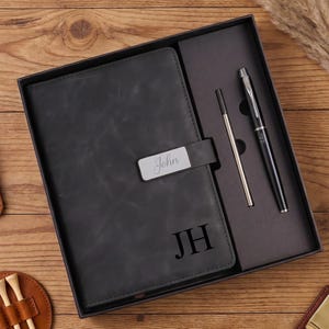 May include: A black notebook with the initials JH embossed on the cover and a silver nameplate reading John, presented in a gift box with a black pen and a refill. Accessories include a brown leather clip, a golf tee holder, and a money clip.