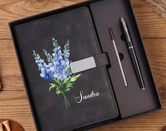 Könnte beinhalten: Ein schwarzes Notizbuch mit einer blauen Blumenillustration und dem in weißer Schreibschrift gedruckten Namen Sandra, ausgestattet mit einem silbernen Metallverschluss. Es liegt in einer schwarzen Geschenkbox mit einem Kugelschreiber.
