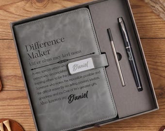 Könnte beinhalten: Ein graues Notizbuch mit dem Text Difference Maker und Daniel, ein schwarzer Stift und eine Metallmine in einer Geschenkbox. Das Set enthält einen braunen Lederriemen, einen runden braunen Lederhalter mit hölzernen Golf-Tees und einen Clip.