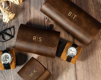 Könnte beinhalten: Drei braune Uhrenetuis aus Leder mit goldgeprägten Initialen B|T, R|S und H|K. Zwei Armbanduhren sind auf beigen Kissen ausgestellt, eine mit silberfarbenem Metallarmband und eine mit dunklem Lederarmband. Eine schwarze Brille ist ebenfalls zu sehen.