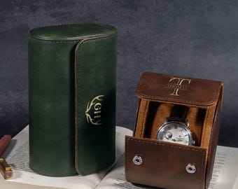 Könnte beinhalten: Ein dunkelgrünes, zylindrisches Uhrenetui aus Leder mit goldenem Geweih-Logo und ein braunes Uhrenetui mit dem Namen THOMAS und einer silbernen Analoguhr darin, beide auf einem offenen Buch neben einem Stift und einem kleinen Metallgegenstand.