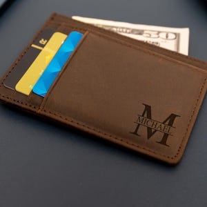 May include: A brown leather wallet with a personalized design, holding several credit cards and a US fifty-dollar bill. The wallet features the name "MICHAEL" and the initial "M" engraved on the front. The wallet is approximately 4 inches wide and 3 inches tall.