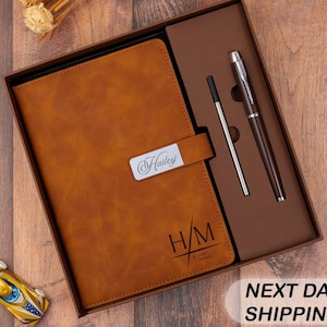 May include: A brown leather journal and pen set in a gift box. The journal has a silver nameplate that reads "Hailey" and initials "H/M". A pen and pen refill are included. The box is on a wooden surface.