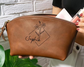Könnte beinhalten: Eine braune Kosmetiktasche aus Leder mit einer schwarzen Strichzeichnung von zwei Blumen in einer Rautenform und dem Namen Sophia in Schreibschrift. Die Tasche verfügt über einen goldfarbenen Reißverschluss und einen braunen Lederanhänger.