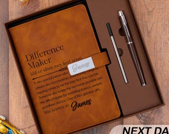 Könnte beinhalten: Ein Notizbuch in hellbrauner Lederoptik mit dem aufgedruckten Text Difference Maker und einer Definition auf dem Einband sowie einem silbernen Namensschild mit dem Namen James. Das Set enthält einen passenden braun-silbernen Kugelschreiber.