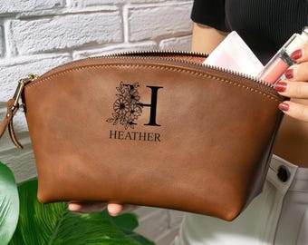 Könnte beinhalten: Eine braune Kosmetiktasche mit Reißverschluss, die mit einem schwarzen Aufdruck eines großen Buchstabens H verziert ist, der von Blumenillustrationen und dem Namen HEATHER darunter begleitet wird. Die Tasche ist halbkreisförmig und matt.