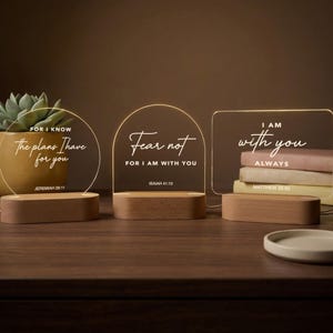 May include: Three illuminated acrylic signs with wooden bases. The signs display inspirational quotes: "For I know the plans I have for you," "Fear not, for I am with you," and "I am with you always." A succulent plant in a yellow pot is visible.