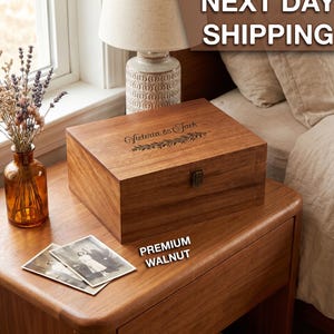 May include: A rectangular wooden keepsake box made of walnut with a hinged lid and a metal latch. The lid features a laser-engraved design with the text Victoria & Jack above a decorative botanical branch motif, showcasing a natural wood grain.