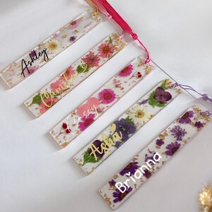 May include: Five rectangular resin bookmarks with dried flowers and gold leaf accents. Each bookmark features a different floral arrangement and a name in gold script: Ashley, Camila, Casey, Asha, and Brianna. Each has a tassel.