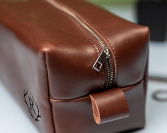 Könnte beinhalten: Eine rechteckige Kulturbeutel aus braunem Leder mit einem silberfarbenen Metallreißverschluss und einer passenden Lederlasche. Die Seite der Tasche ist mit einem dekorativen Motiv geprägt, das den Buchstaben R zwischen zwei Hirschgeweihen zeigt.