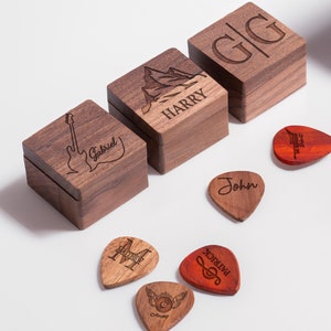 May include: Six wooden boxes with engraved names and a mountain design.  The boxes are arranged in a row on a white surface.  There are also six guitar picks with engraved designs, including a musical note, a record player, and a mountain.