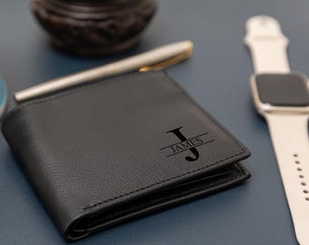 Könnte beinhalten: Ein schwarzes Leder-Brieftaschen-Faltmodell mit einer personalisierten Gravur des Buchstabens J, wobei der Name JAMES horizontal über den Buchstaben zentriert ist. Die Brieftasche hat sichtbare Nähte an den Kanten und eine glatte, matte Oberfläche.
