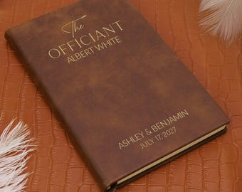 Könnte beinhalten: Ein braunes Notizbuch in Lederoptik mit goldfarbenem Text, der The Officiant Albert White und Ashley & Benjamin July 17, 2027 lautet. Das rechteckige Journal hat eine glatte, matte Oberfläche und ein schmales, dunkelbraunes Lesebändchen unten.
