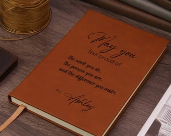 Könnte beinhalten: Ein Notizbuch mit beigem Ledereinband und einer schwarzen Gravur mit dem Text: May you feel proud of the work you do, the person you are, and the difference you make. For Ashley. Das Notizbuch enthält ein passendes beiges Lesebändchen.