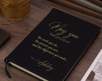 Könnte beinhalten: Ein schwarzes Notizbuch mit goldener Schrift auf dem Einband: May you feel proud of the work you do, the person you are, and the difference you make. For Ashley. Das Notizbuch verfügt über ein schwarzes Lesebändchen und eine strukturierte Oberfläche.