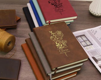 Könnte beinhalten: Ein Stapel Notizbücher mit Kunstledereinbänden in verschiedenen Farben, darunter Kastanienbraun, Blaugrün, Blau, Weiß, Schwarz, Braun, Grau und Hellbraun. Das oberste kastanienbraune Notizbuch zeigt ein schwarzes Blumenmotiv und den Namen Ashley. Das oberste braune Notizbuch zeigt ein goldenes Blumenmotiv und den Namen Olivia.
